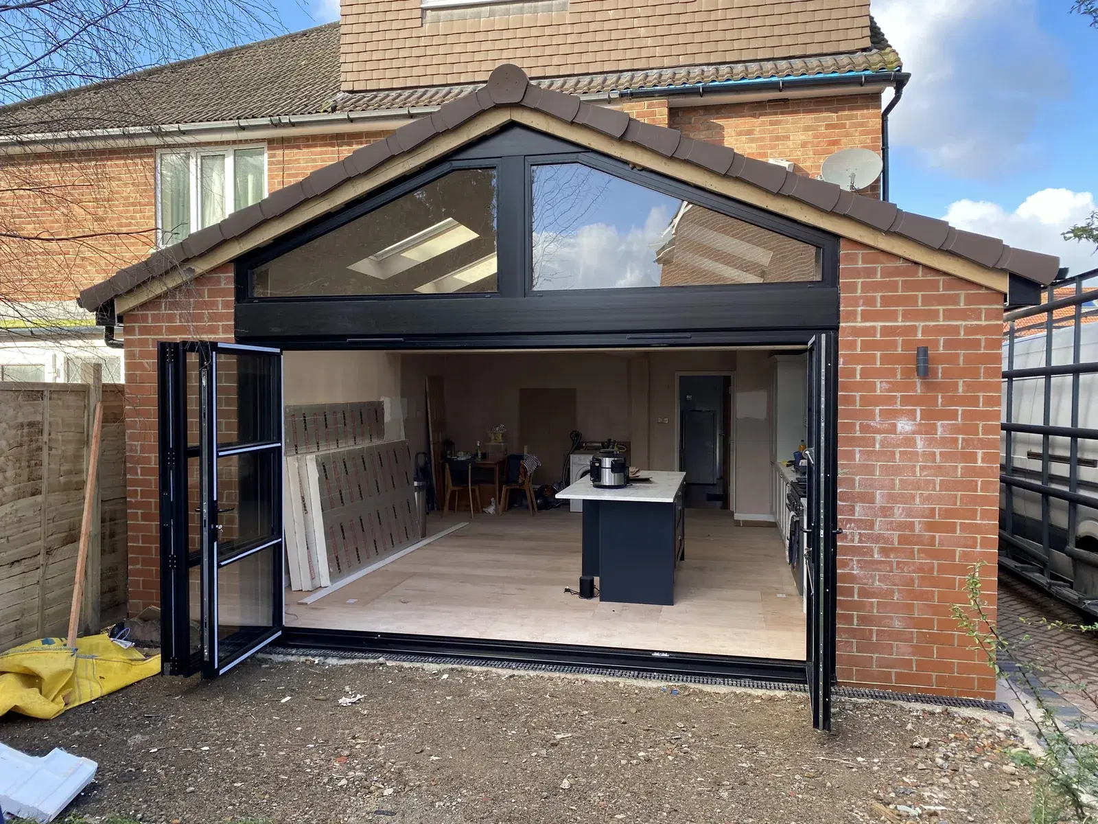 Black bi-folding doors folded open on a red-brick extension, with an apex gable window above and a kitchen visible inside.