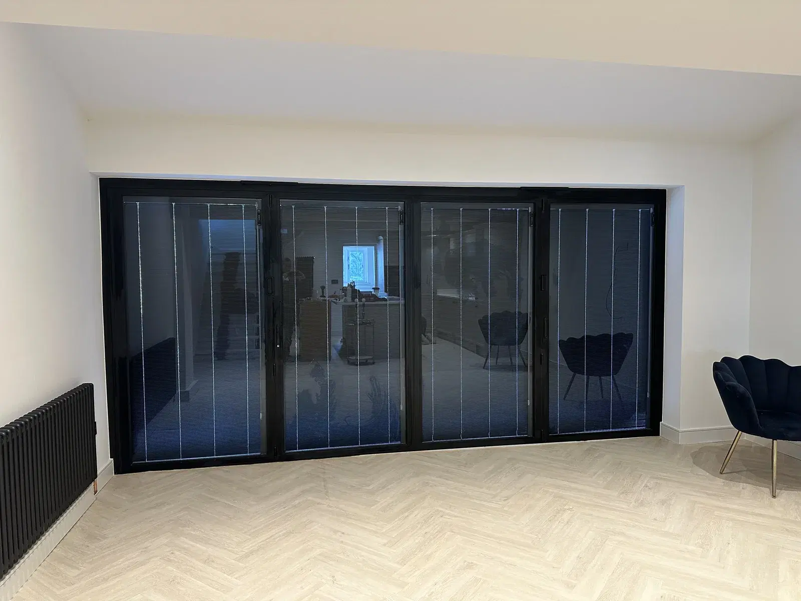 Black bi-folding doors with integral blinds, seen from inside a room with a herringbone floor.