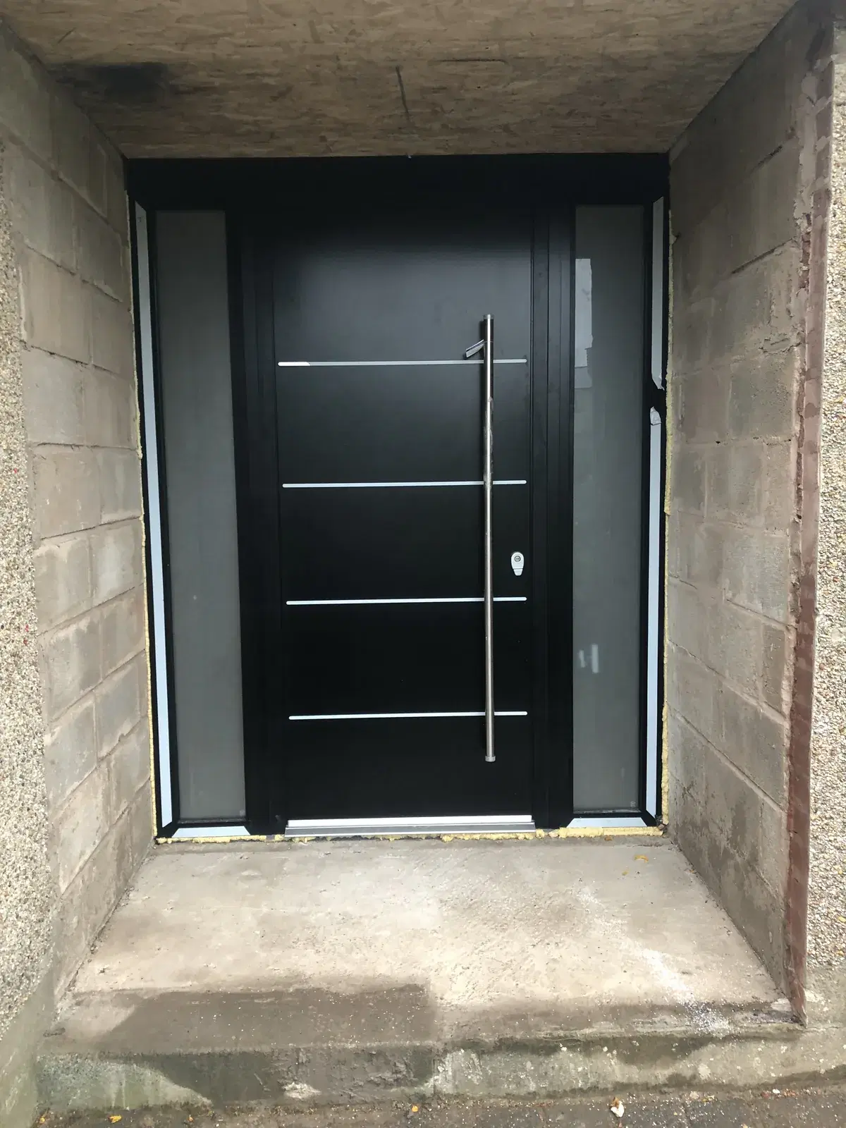 A black entrance door with horizontal chrome inlays, flanked by two frosted side panels.