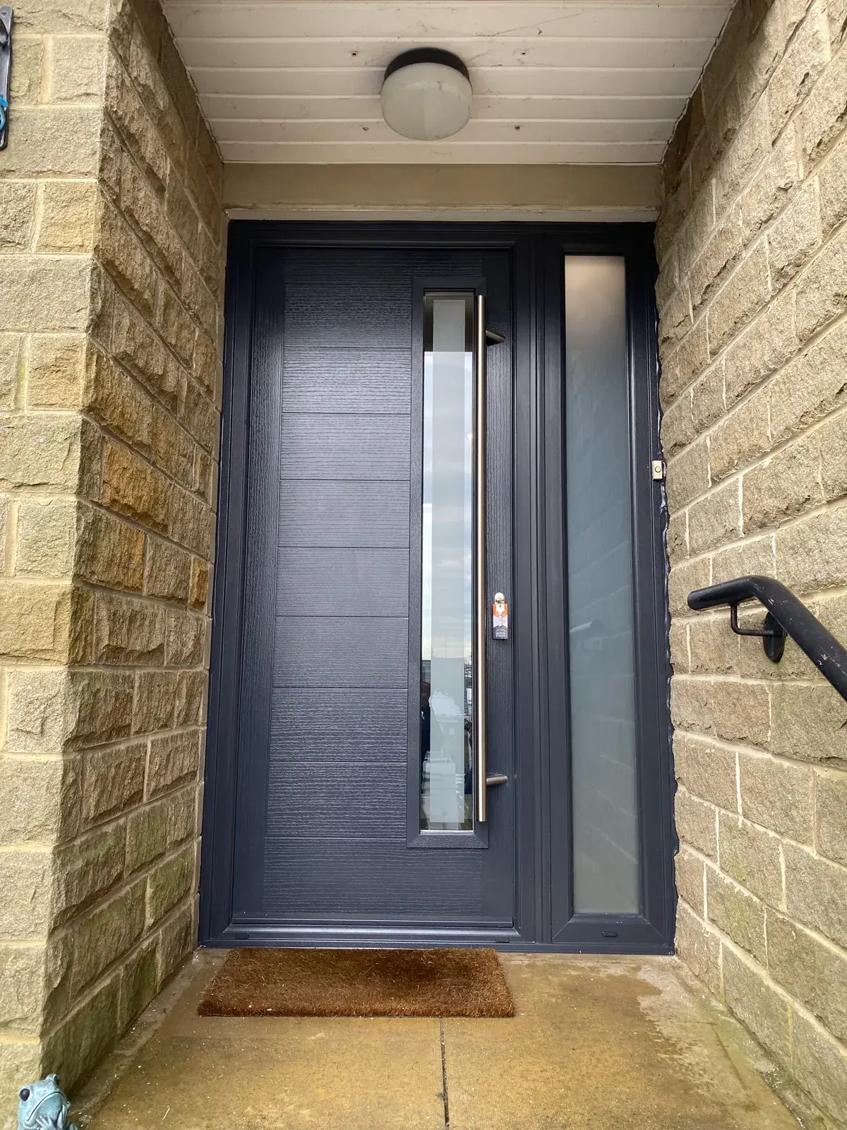 An anthracite composite door with a long bar handle, a vertical glazed panel and a frosted side light.