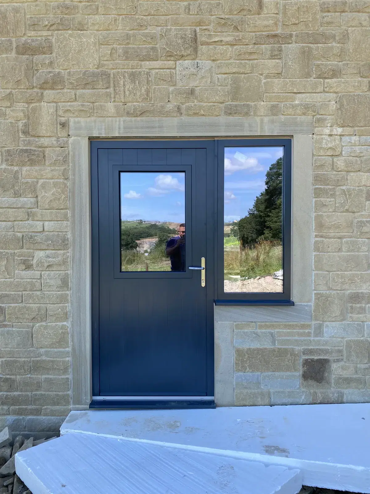 A dark blue stable-style door with a glazed upper panel and a matching side window, in a stone-built house.