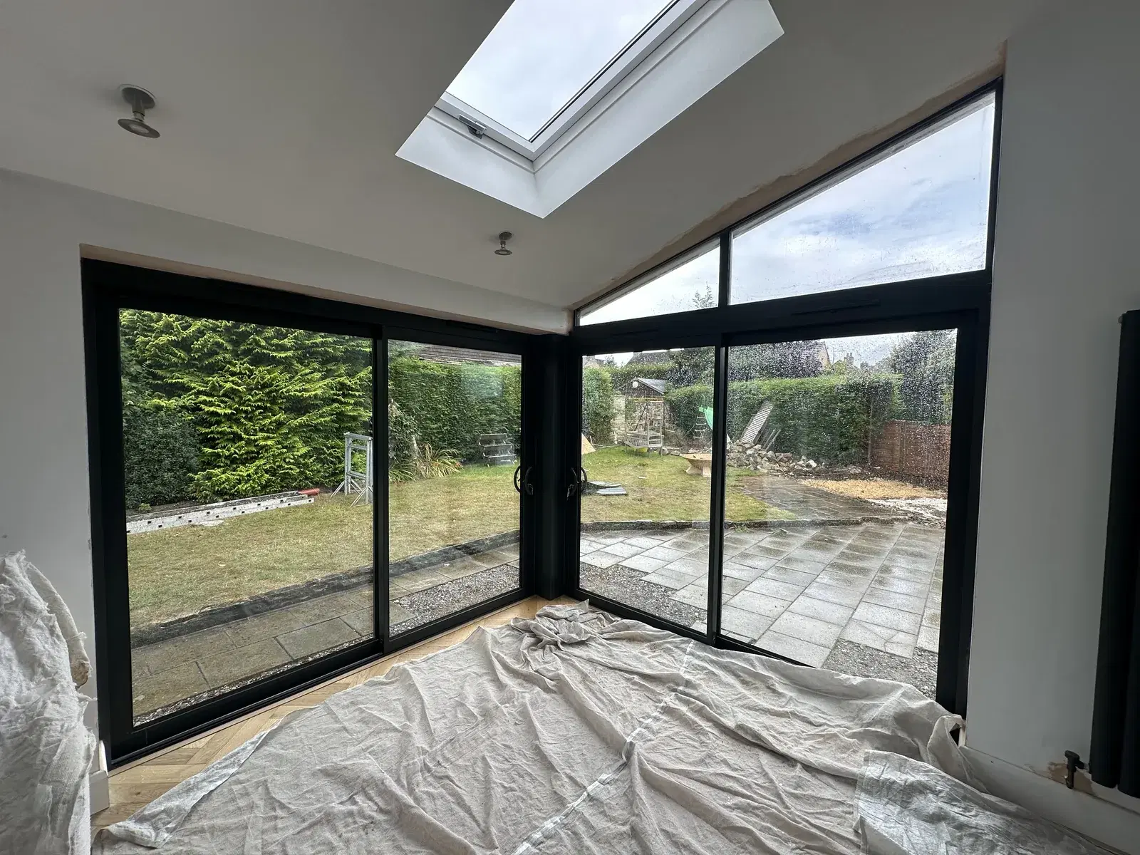 Black sliding doors wrapping a corner of a new extension, with a roof light above and dust sheets still down.