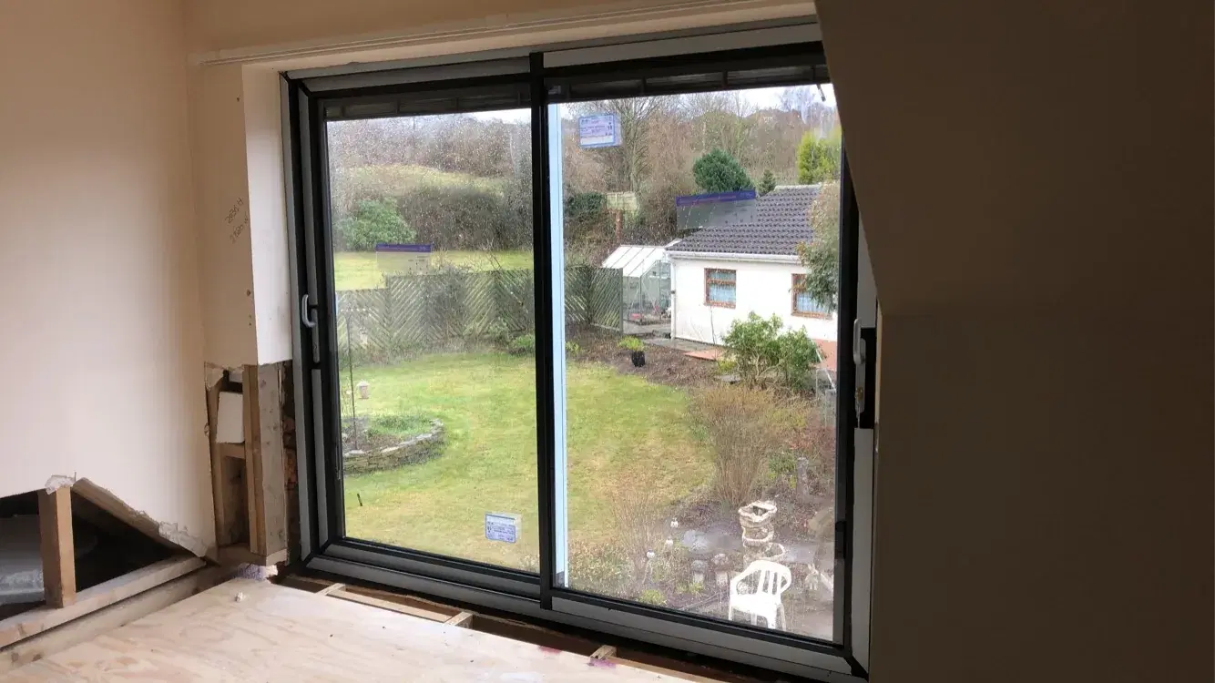A dark grey sliding door seen from inside a room mid-renovation, overlooking a long garden.