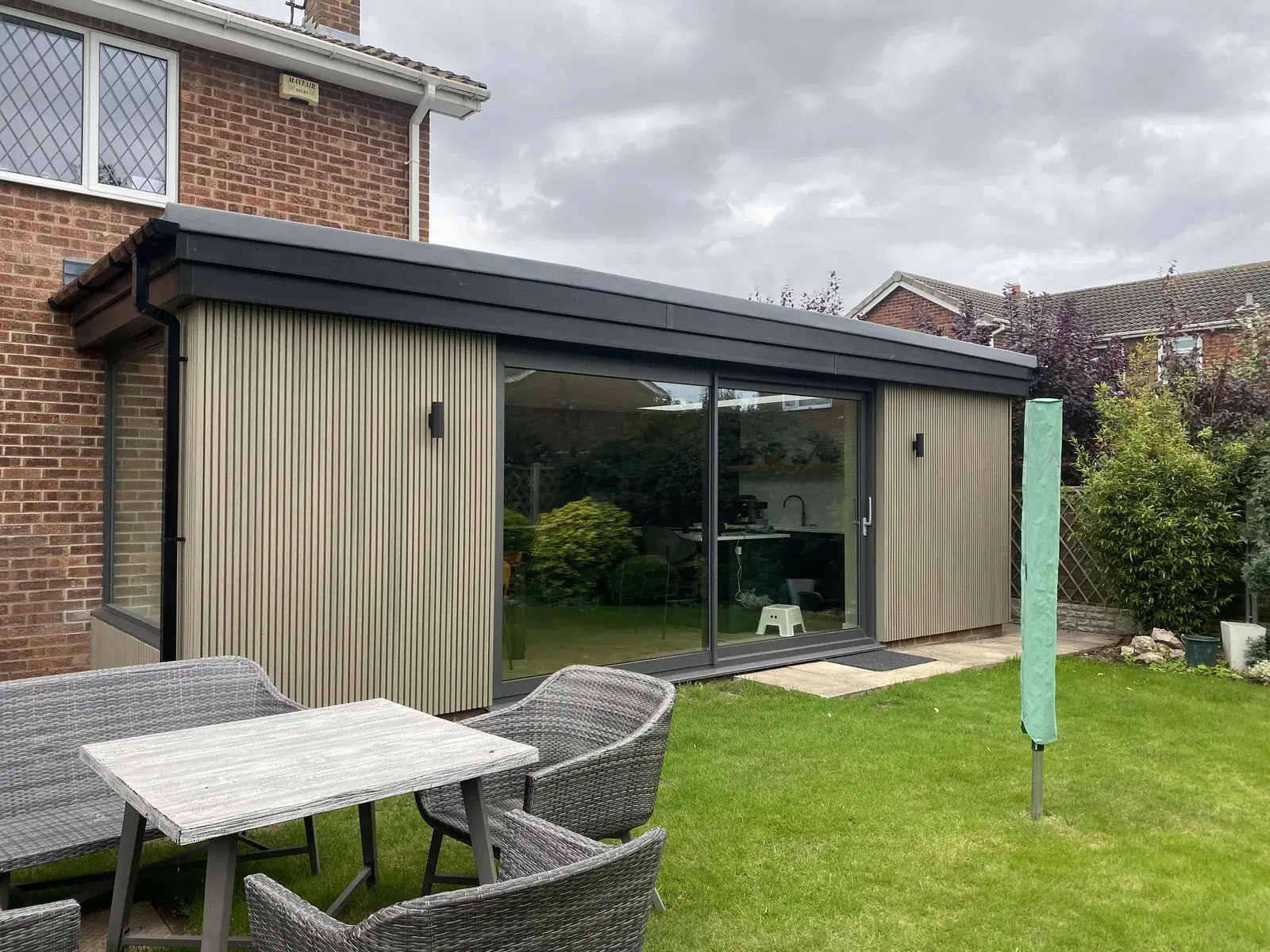 Anthracite sliding patio doors set between fluted cladding on a garden room, with a rattan table on the lawn.
